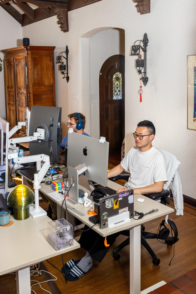 Two people work at desks with large computer monitors in a room with wooden floors, white walls, and vintage decor including a wooden armoire and wall sconces.